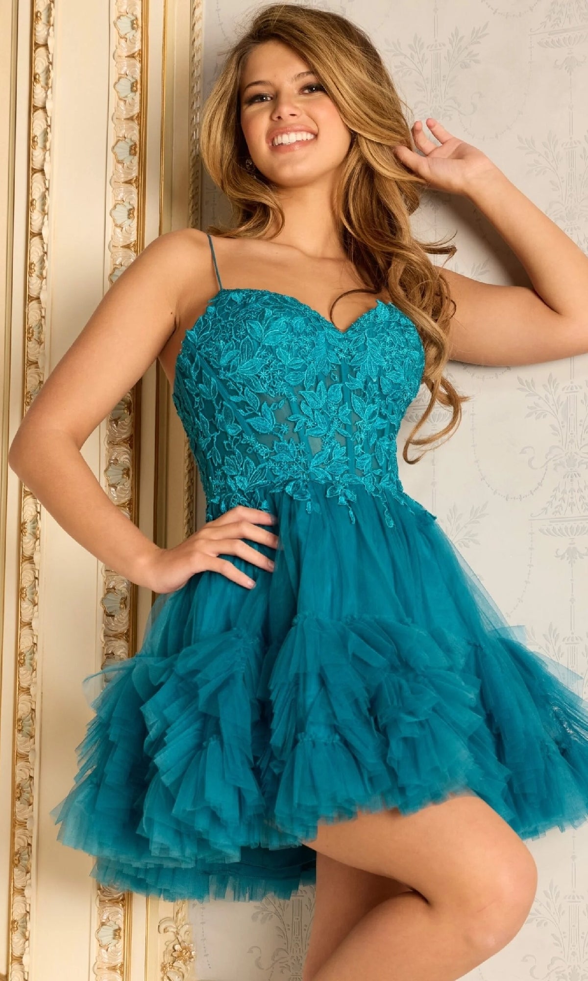 A woman smiles indoors, posing with one hand in her hair while wearing the Jovani 45335 short homecoming dress—a teal, lace-embroidered design featuring spaghetti straps and a ruffled tulle skirt.